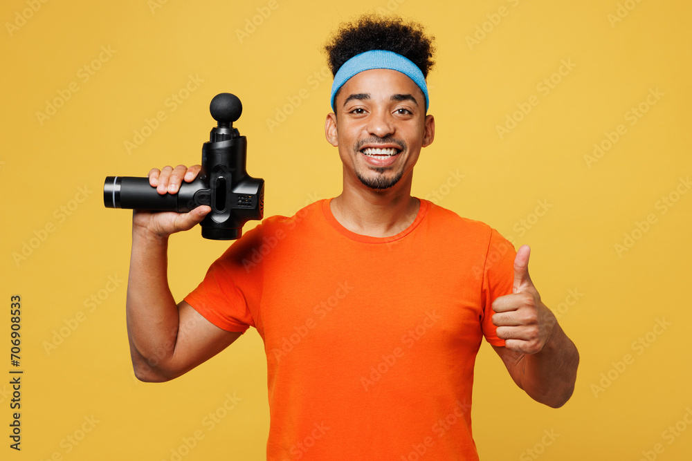 Young fitness trainer instructor sporty man sportsman wear orange t-shirt hold percussion massager show thumb up training in home gym isolated on plain yellow background Workout sport fit abs concept Young fitness trainer instructor sporty man sportsman wear orange t-shirt hold percussion massager show thumb up training in home gym isolated on plain yellow background Workout sport fit abs concept