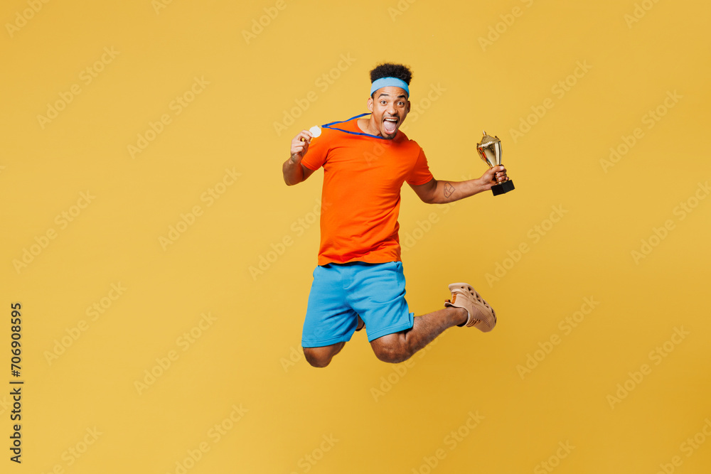 Full body young fitness trainer instructor sporty man sportsman wear orange t-shirt jump high golden medal champion cup award in gym isolated on plain yellow background. Workout sport fit abs concept. Full body young fitness trainer instructor sporty man sportsman wear orange t-shirt jump high golden medal champion cup award in gym isolated on plain yellow background. Workout sport fit abs concept.