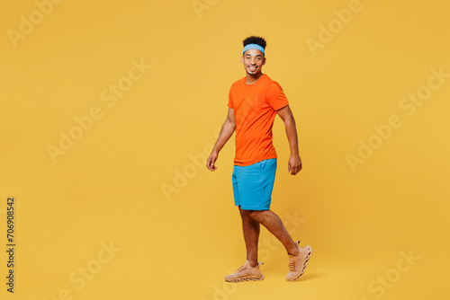 Full body side view happy young fitness trainer sporty man sportsman wear orange t-shirt walk go look camera spend time in home gym isolated on plain yellow background. Workout sport fit abs concept.