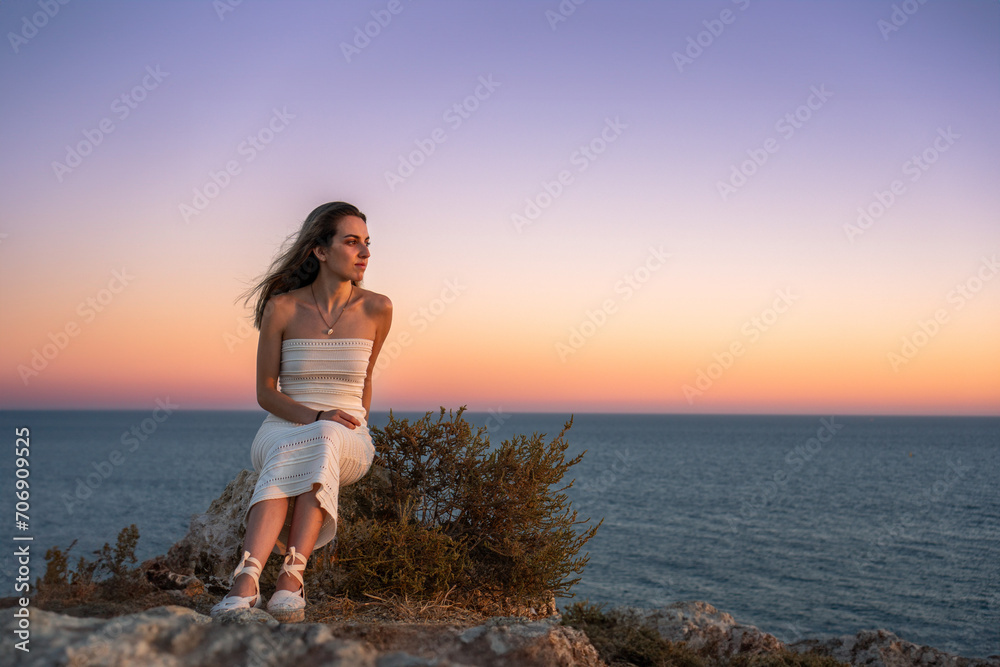 Stylish young woman sitting on rocky seashore and looking away