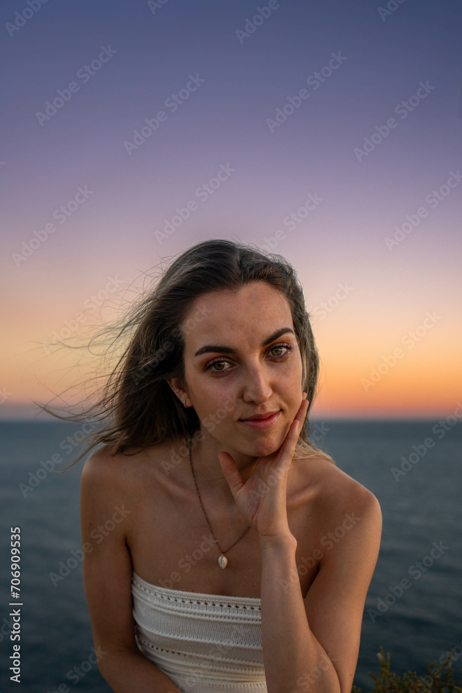 Vertical CloseUp portrait Capture of a Young Woman in a White Dress, with a Gold Necklace, sitting by the Ocean at Sunset, Hair Styled in Loose Waves, Set Against a Backdrop of a Sky 