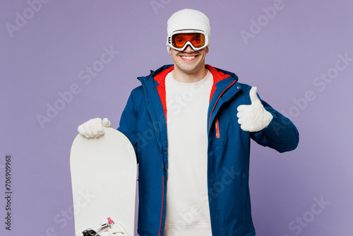 Happy smiling fun cool man he wear warm blue windbreaker jacket ski goggles mask hat hold snowboard show thumb up spend extreme weekend winter season in mountains isolated on plain purple background.