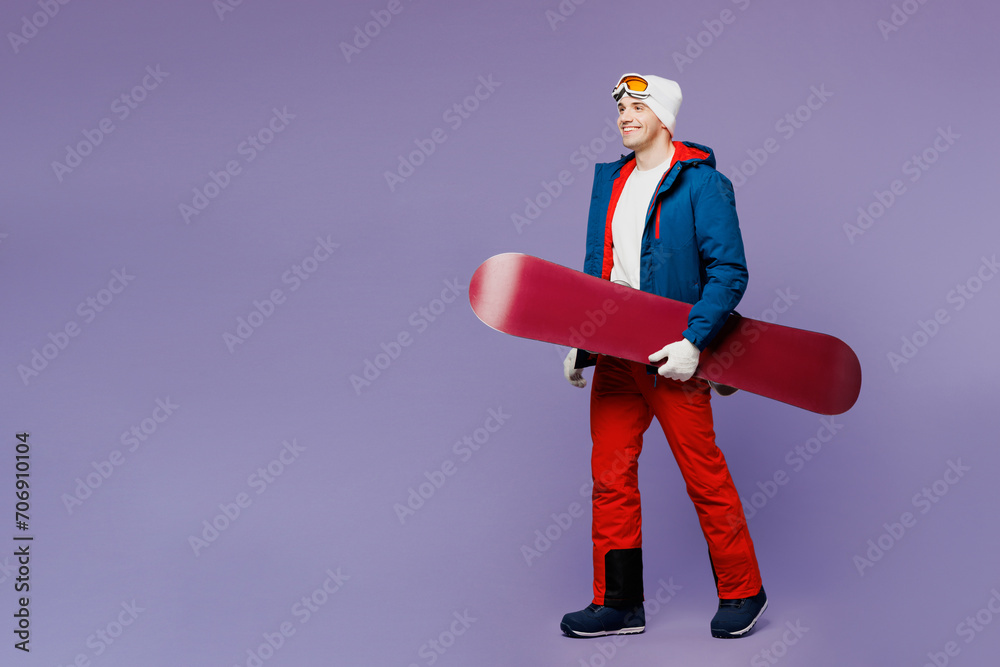 Full body side view smiling happy fun man wear blue windbreaker jacket ski goggles mask hat hold snowboard go walk spend extreme weekend winter season in mountains isolated on plain purple background. Full body side view smiling happy fun man wear blue windbreaker jacket ski goggles mask hat hold snowboard go walk spend extreme weekend winter season in mountains isolated on plain purple background.