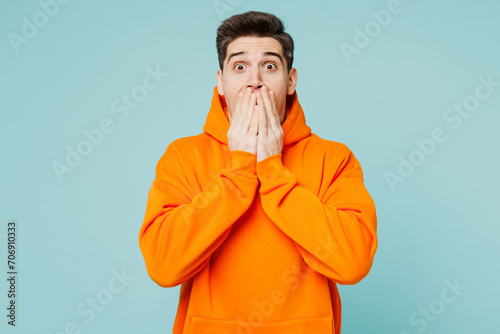 Young shocked surprised happy man he wears orange hoody casual clothes cover mouth with hand look camera isolated on plain pastel light blue cyan color background studio portrait. Lifestyle concept.