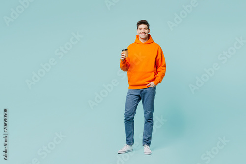 Full body young man he wears orange hoody casual clothes hold takeaway delivery craft paper brown cup coffee to go isolated on plain pastel light blue cyan color background studio. Lifestyle concept.