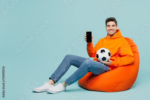 Full body young man fan in casual clothes cheer up support football sport team hold soccer ball watch tv live stream sit bag chair use blank screen mobile cell phone isolated on plain blue background.
