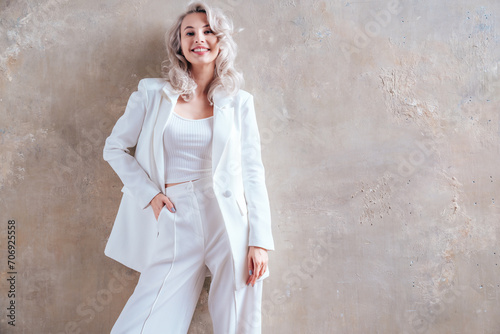 Portrait of young beautiful blond woman wearing nice trendy white suit. Fashion model posing in studio. Fashionable female posing near grey wall. Cheerful and happy