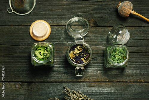Dry aromatic herbs in glass jars on a wooden table, top view.