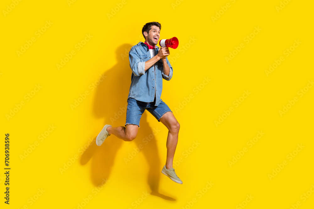 Full size photo of cheerful guy wear jeans jacket jump look empty space announcing sale in loudspeaker isolated on yellow color background