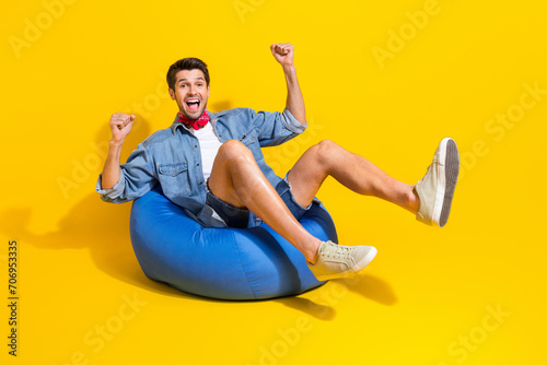 Full body photo of overjoyed man dressed denim shirt red scarf sit on bean bag win lottery isolated on vibrant yellow color background