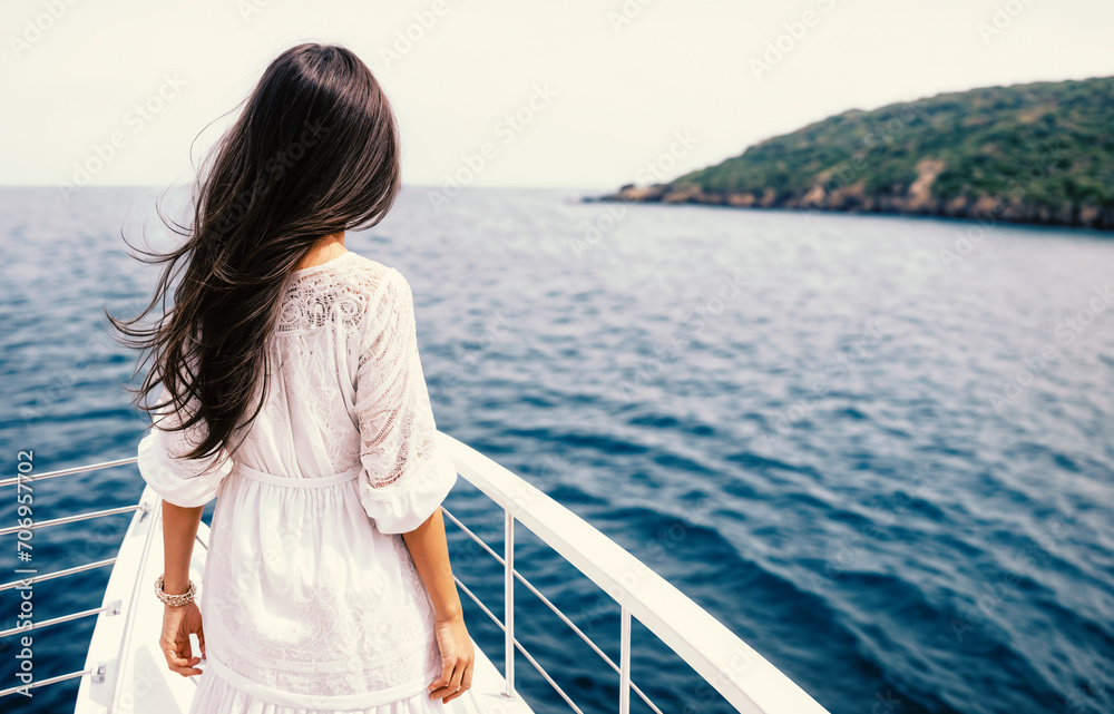 A beautiful woman with long brown hair stands on the deck of a yacht in a white dress with lace and looks at the seascape. Back view. 