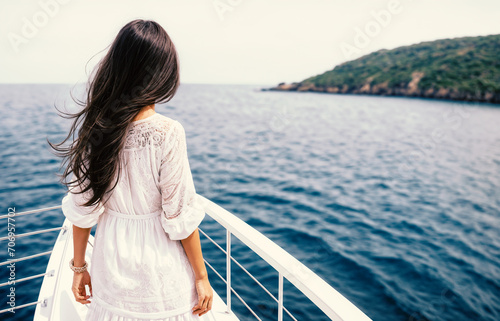 A beautiful woman with long brown hair stands on the deck of a yacht in a white dress with lace and looks at the seascape. Back view. 