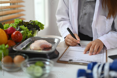 Cropped shot of female dietitian sitting at desk with different healthy products and writing diet plan