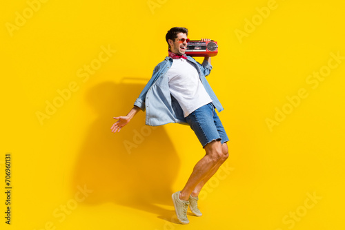 Full size photo of eccentric guy wear jeans jacket in sunglass boombox on shoulder look empty space isolated on yellow color background