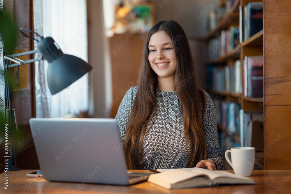 Fototapeta premium Smiling girl having online lesson via the Internet. Cheerful female student in university library, preparing assignment