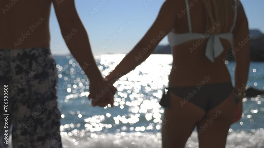 Beautiful pair holding hands of each other and walking to sea on sunny day. Young couple spending time together during summer travel. Sun light reflection at the water surface at background. Close up Beautiful pair holding hands of each other and walking to sea on sunny day. Young couple spending time together during summer travel. Sun light reflection at the water surface at background. Close up