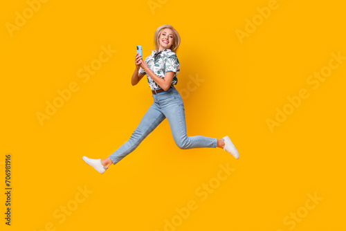 Full length photo of optimistic woman wear blouse denim pants jumping with smartphone in hands isolated on yellow color background