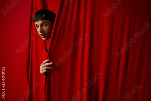 playful young man in vibrant shirt hiding behind red curtain while playing hide and seek, peeking photo