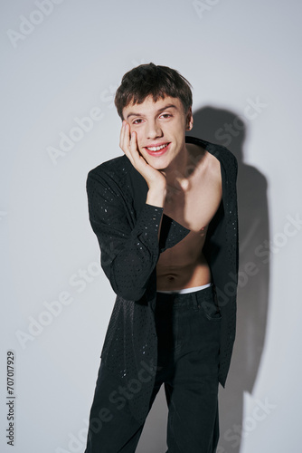 cheerful young man in dotted black shirt and jeans posing with hand near cheek on grey background