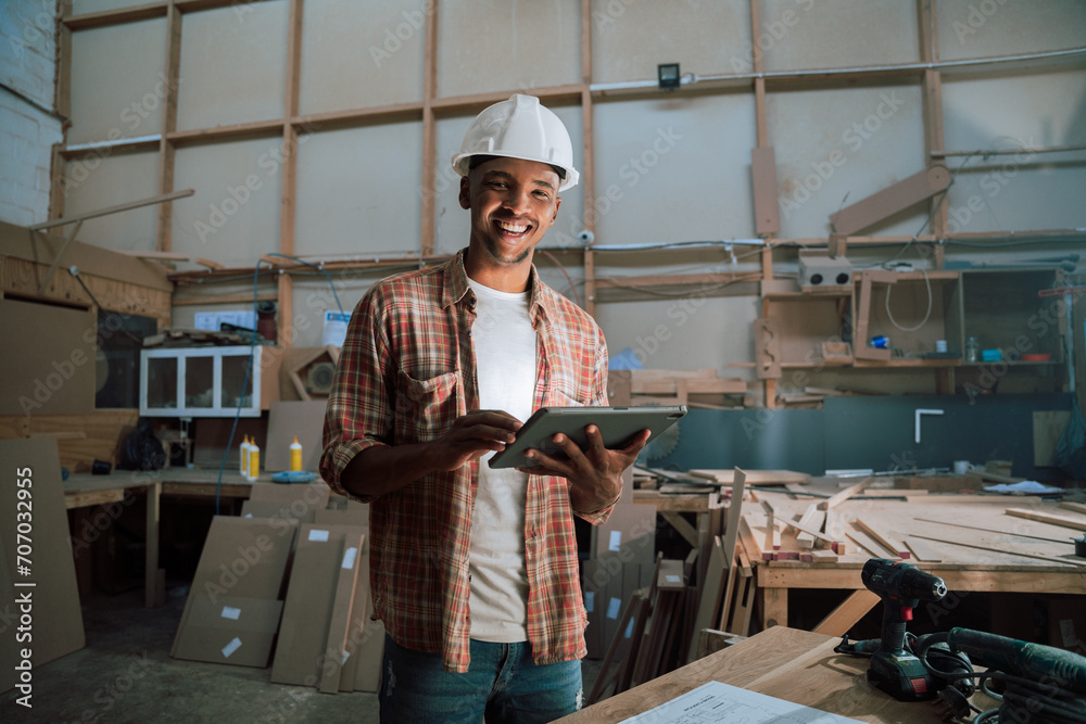 Smiling African American with hardhat smiles while using tablet in carpentry warehouse Smiling African American with hardhat smiles while using tablet in carpentry warehouse
