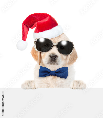 Cute Golden retriever puppy wearing sunglasses, red santa hat and tie bow looks above  empty white banner. isolated on white background © Ermolaev Alexandr
