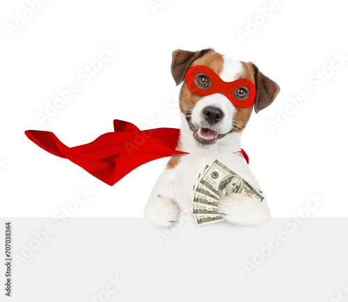 Funny jack russell terrier puppy wearing superhero costume looking above empty white banner and shows dollars usa. Isolated on white background