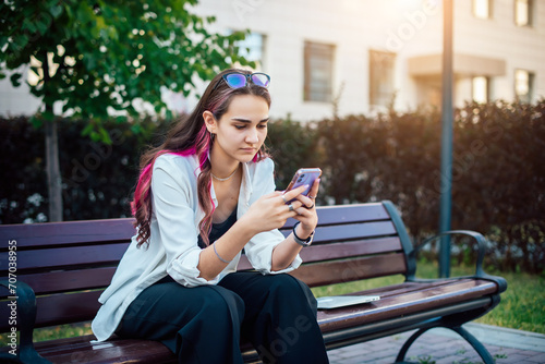Portrait shot of a young brunette woman messaging on mobile phone while sitting on the bench outdoors. Beautiful female person using smartphone outside.