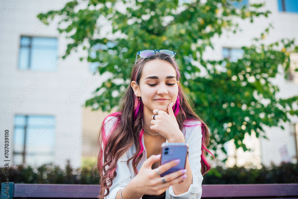 Portrait shot of a young brunette woman messaging on mobile phone while sitting on the bench outdoors. Beautiful female person using smartphone outside. Portrait shot of a young brunette woman messaging on mobile phone while sitting on the bench outdoors. Beautiful female person using smartphone outside.