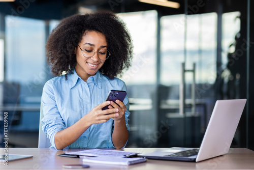 Businesswoman at workplace inside office  employee uses app on phone  woman happily smiles browsing social media and reading text message.