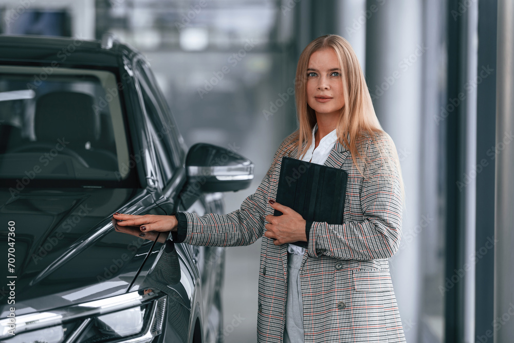 Fototapeta premium With tablet in hands. Woman in formal clothes is in the car dealership
