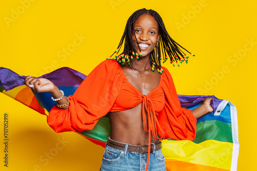 African american young smiling girl with braids, toothy smiles with pride rainbow flag in motion at studio isolated over yellow background. Trans or bisexual concept.