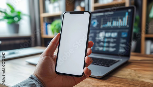 Mockup white blank screen of mobile phone in male hand in front of blurred laptop with financial charts.