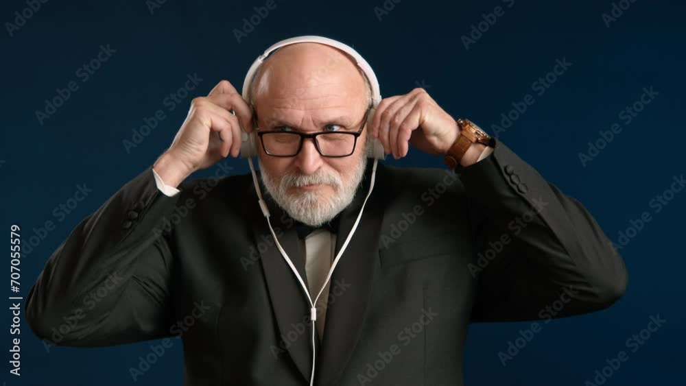 Distinguished man in a black tuxedo, putting on white headphones and enchantingly listening to music. Classic glasses and a bow tie around his neck give him an elegant style. Background is dark blue. Distinguished man in a black tuxedo, putting on white headphones and enchantingly listening to music. Classic glasses and a bow tie around his neck give him an elegant style. Background is dark blue.