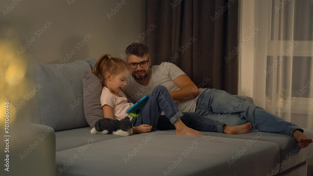 Happy family, father and little girl watch digital tablet sitting on sofa at home in evening. Leisure. Computer games, surfing internet, social media Happy family, father and little girl watch digital tablet sitting on sofa at home in evening. Leisure. Computer games, surfing internet, social media