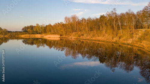 This serene image captures the calm waters of a river during the golden hour, with the warm light of the setting sun illuminating the trees and grasses along the banks. The trees, stripped of their