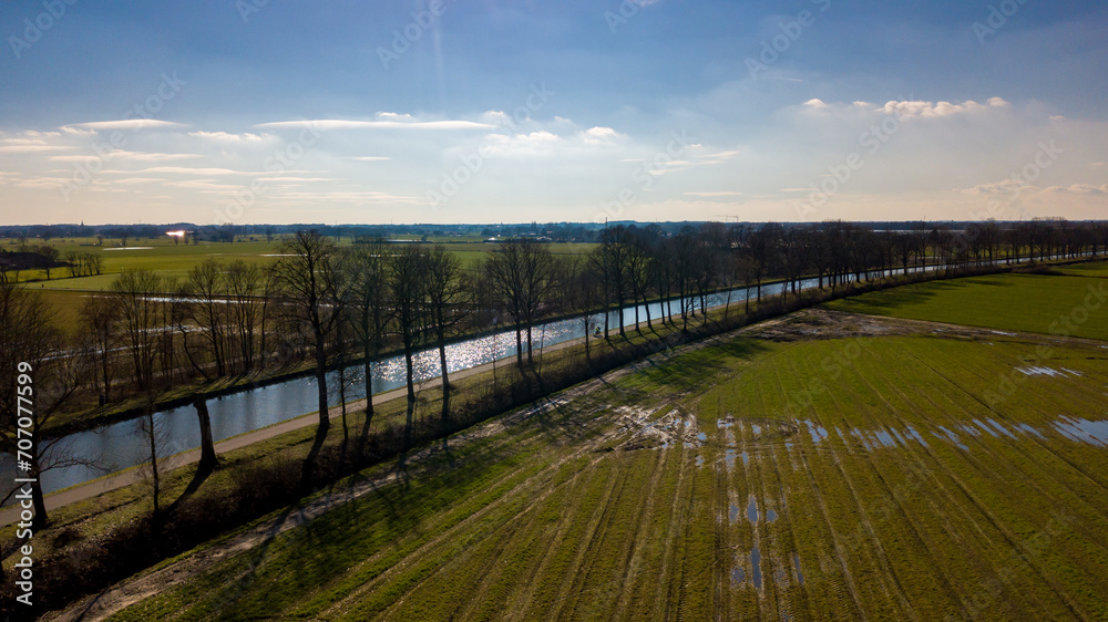 The photograph encapsulates a serene rural landscape, taken from an aerial perspective. It features a glistening river that catches the rays of the sun, bordered by a row of trees casting elongated