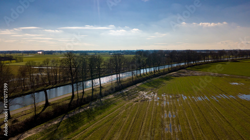 The photograph encapsulates a serene rural landscape, taken from an aerial perspective. It features a glistening river that catches the rays of the sun, bordered by a row of trees casting elongated