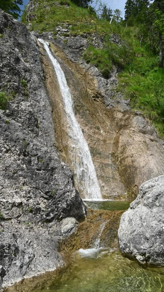 Vertical shot of a waterfall next to a steep meadow in the mountains, with a pond in the foreground, direct sunlight Vertical shot of a waterfall next to a steep meadow in the mountains, with a pond in the foreground, direct sunlight