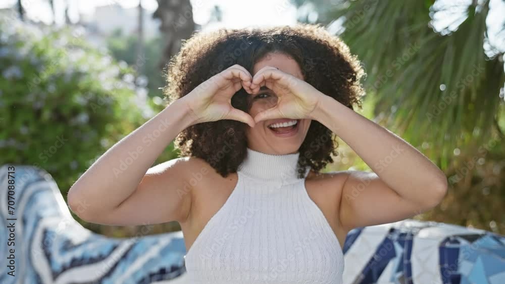 Young hispanic woman with curly hair making heart shape with hands, smiling in a sunny outdoor park setting. Young hispanic woman with curly hair making heart shape with hands, smiling in a sunny outdoor park setting.