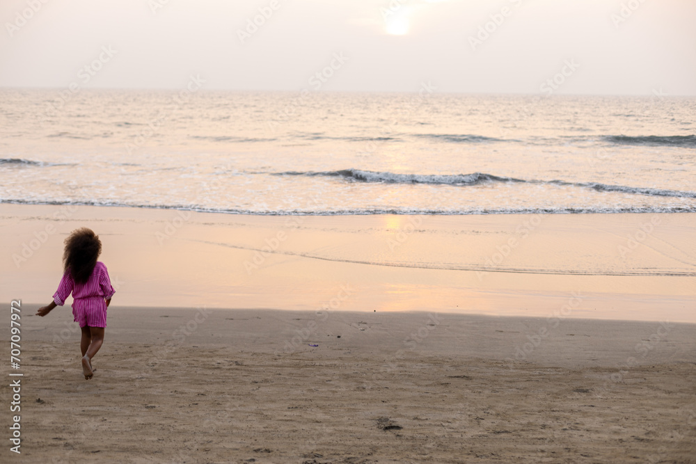 Unrecognizable girl with loose hair and dark skin runs along sandy beach to sea at sunset. Layout. Unrecognizable girl with loose hair and dark skin runs along sandy beach to sea at sunset. Layout.