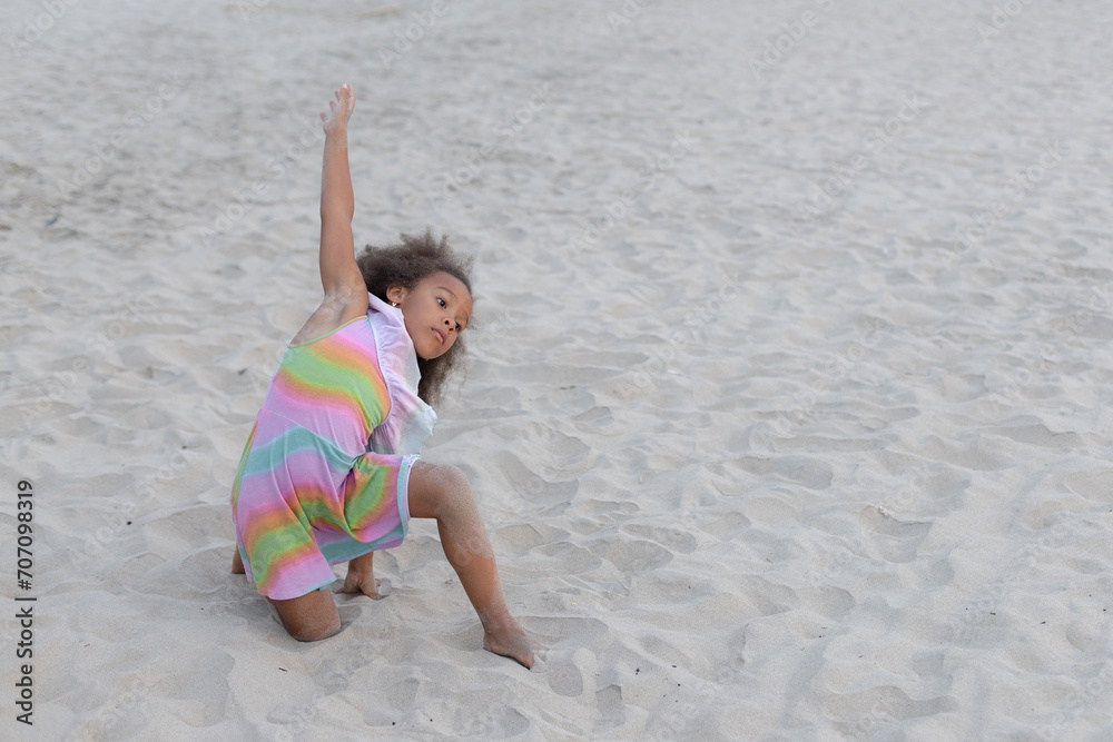 Children's yoga. Little girl with dark skin is sitting on sand on beach, doing yoga exercise.