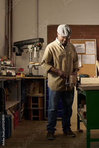 Vertical portrait of African American senior man as skilled carpenter standing in sunlit workshop
