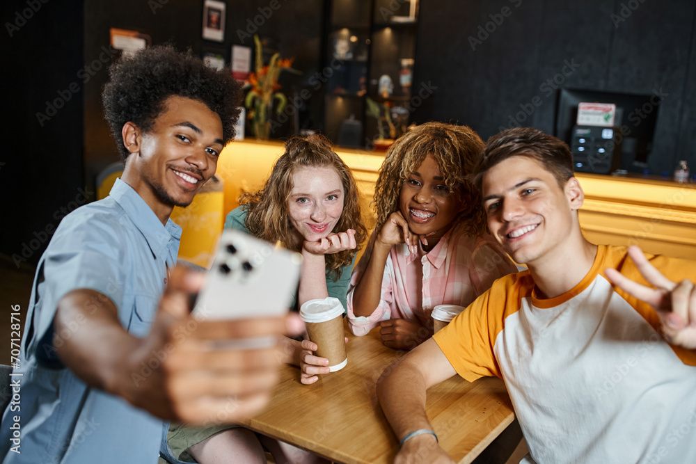 Fototapeta premium cheerful african american man taking selfie with stylish multiethnic friends in lounge of hostel