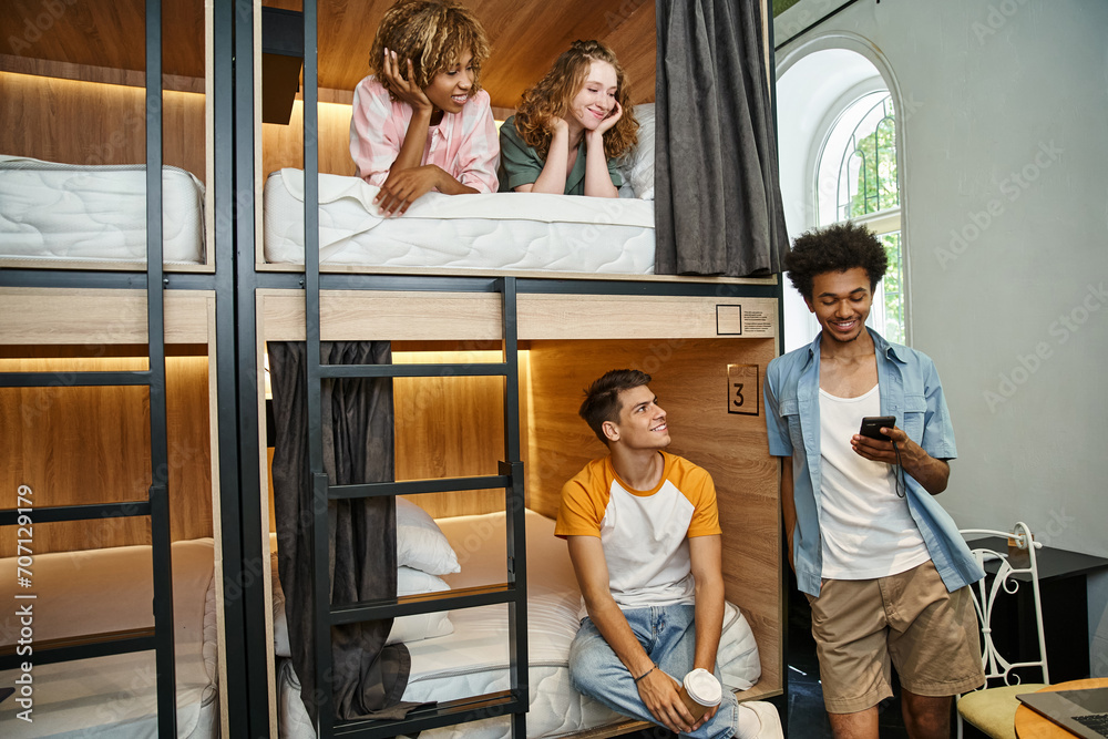 smiling african american man using smartphone near multiethnic mates on double-decker beds in hostel