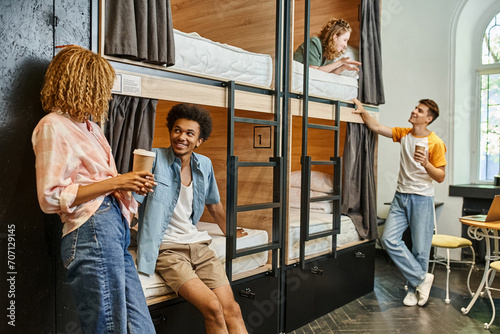 cheerful multicultural roommates with coffee to go talking near double-decker beds in youth hostel photo