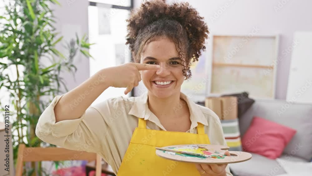 Cheerful hispanic woman artist with curly hair, playfully pointing finger to her fresh, natural beauty face and nose, while sitting joyfully in the studio. Cheerful hispanic woman artist with curly hair, playfully pointing finger to her fresh, natural beauty face and nose, while sitting joyfully in the studio.