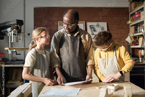 Black senior carpenter teaching two kids woodworking in small workshop and showing them plans