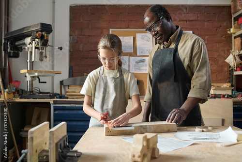 Waist up portrait of young girl measuring wood in workshop with senior carpenter watching