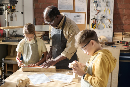 Portrait of senior craftsman teaching children carpentry in workshop lit by sunlight, copy space