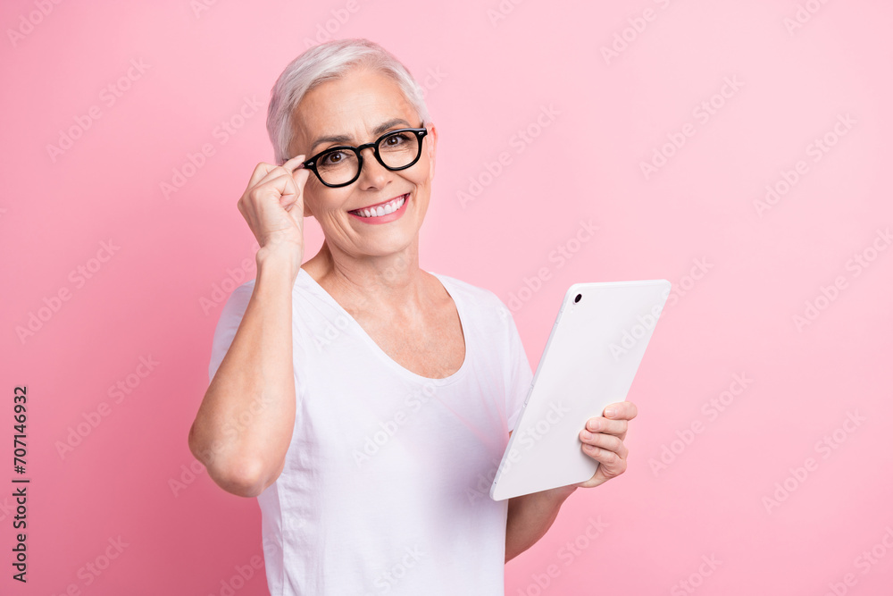 Photo of senior professional woman with short hairstyle wear white t-shirt touch glasses holding tablet isolated on pink background Photo of senior professional woman with short hairstyle wear white t-shirt touch glasses holding tablet isolated on pink background